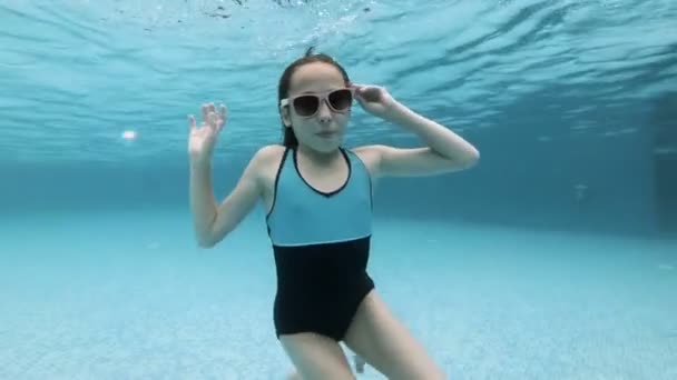 Una bella bambina posa sott'acqua in una piscina con i capelli sciolti in una giornata di sole in occhiali da sole rosa su uno sfondo blu. Guarda la telecamera e agita le mani. Moda. Al rallentatore. 4K . — Video Stock