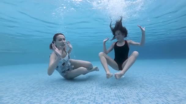 Dos niñas, hermanas de diferentes edades, juegan bajo el agua en la piscina. Se sumergen y se sientan en el fondo de la piscina, miran a la cámara, saludan y sonríen. Retrato. En cámara lenta. 4K . — Vídeo de stock