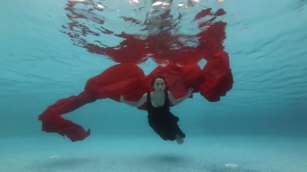 Een jong mooi meisje in een zwarte jurk zwemt en speelt onderwater in een zwembad met een lange rode doek op een blauwe achtergrond. Modeportret. Kunstvideografie. Langzame beweging. 4k. — Stockvideo