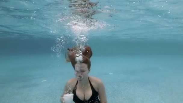 Сексуальная молодая блондинка с длинными волосами ныряет и позирует под водой в бассейне с белой чашкой и блюдцем в руке в черном купальнике. Она пускала пузыри и смотрела в камеру. Медленное движение. 4K . — стоковое видео