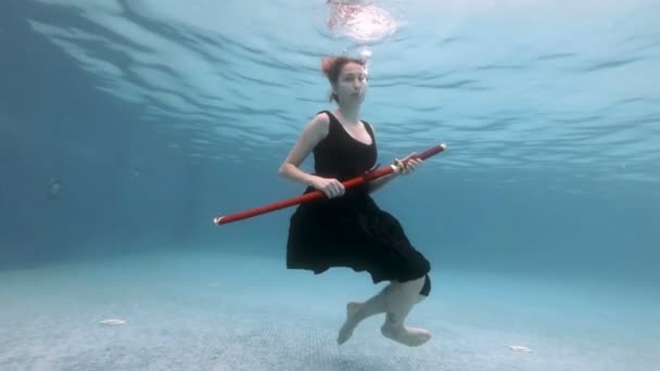 Uma jovem afunda-se no fundo da piscina com uma espada samurai vermelha na mão com um vestido preto. Ela tira a lâmina da bainha e mostra-a à câmara. Retrato. Arte. Movimento lento. 4K . — Vídeo de Stock