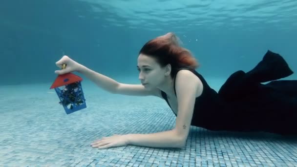 Une belle jeune fille avec un cadeau de Noël à la main rampe sous l'eau au fond de la piscine dans une robe noire. Il s'éloigne du fond et flotte jusqu'à la surface. Au ralenti. 4K . — Video