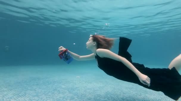 Beautiful young girl swimming underwater in a pool in a black dress with a Christmas gift in her hand. She holds the toy at arm's length and releases bubbles. Christmas. Portrait. Slow motion. 4K. — Stock Video