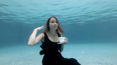 Siyah elbiseli güzel bir kız havuzun dibinde su altında beyaz bir kupadan su içiyor. Kameraya bakar ve gülümser. Moda portresi. Yaratıcı videografi. Ağır çekim. 4k.