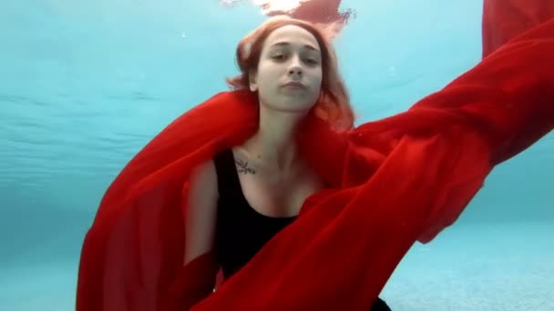 Portrait of a young girl with a red cloth under water. She emerges from the bottom to the surface holding the red cloth in her hand and looks into the camera. Close-up. Bottom view. Slow motion. 4K. — Stock Video