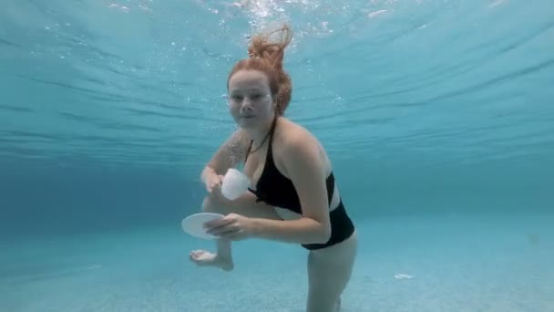 Una hermosa joven con el pelo largo bucea y posa bajo el agua con una taza blanca y un platillo en la mano en un traje de baño negro. Ella mira a la cámara y los POP arriba. En cámara lenta. 4K . — Vídeo de stock