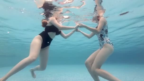 Two girls in red Santa hats hold hands and dive under the water. They pose for the camera and smile near the bottom of the pool holding hands. Portrait. Close-up. Slow motion. 4K. — Stock Video