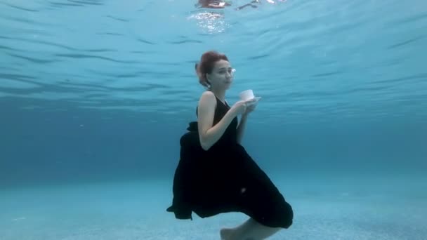 Une jeune fille souriante descend lentement et s'assoit au fond de la piscine dans une robe noire avec une tasse blanche à la main. Elle soufflait des bulles, ajustait sa robe et souriait. Portrait. Au ralenti. 4K . — Video