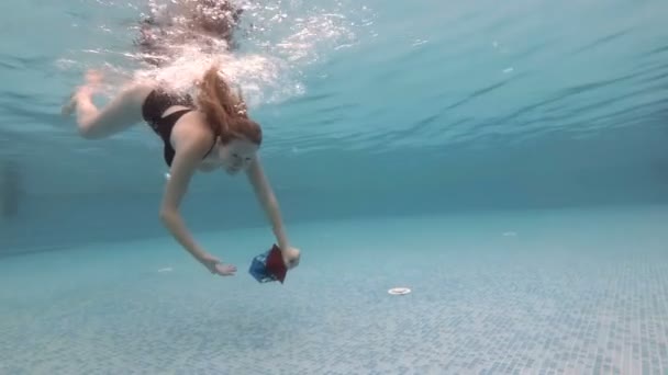 Sexy joven con el pelo largo que fluye inmersiones y nada bajo el agua en la piscina con un regalo de Navidad en su mano extendida en un traje de baño negro y deja burbujas. En cámara lenta. 4K . — Vídeos de Stock