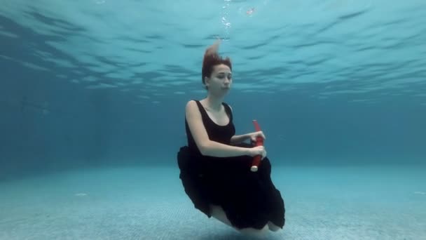 Surrealistyczna dziewczyna powoli tonie pod wodą na dnie basenu w czarnej sukience z czerwonym samurajskim mieczem w ręku. Wyciąga miecz, pokazuje go i bawi się nim. Portret. Zwolniony ruch. — Wideo stockowe