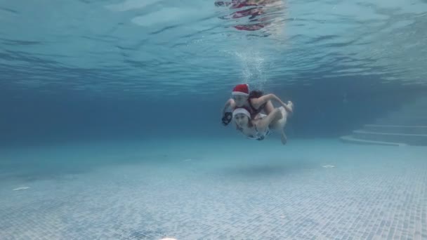 Zwei junge Mädchen in roten Weihnachtsmützen schwimmen gemeinsam unter dem Wasser im Pool. Ein kleines Mädchen schwebt auf dem Rücken ihrer älteren Schwester. Sie lächeln und blicken in die Kamera. Ansicht von unten. Zeitlupe. 4k. — Stockvideo