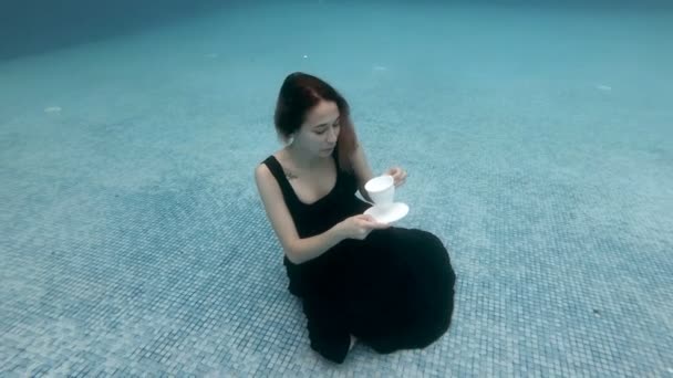 Aranyos játékos lány mosolyog, és nézi a kamerát ül a víz alatt a medence alján egy fekete ruhát egy fehér csésze a kezében. Aztán lassan lebeg a felszínre. Fogantatás. Lassú mozgás.. — Stock videók
