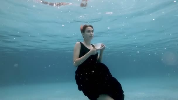 Красивая девушка в черном платье медленно тонет под водой с белой чашкой и блюдцем в руках в облаке пузырьков. Она смотрит в камеру и выпрямляет волосы. Медленное движение. 4K . — стоковое видео