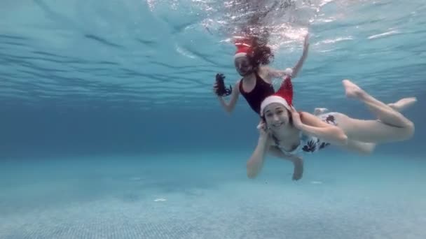 Malá holčička a její starší sestra v červených kloboucích Santa plavou pod vodou ve veřejném bazénu. Usmějí se a dívají se do kamery. Neobvyklé Vánoce. Portrét. Zpomal. 4k. — Stock video