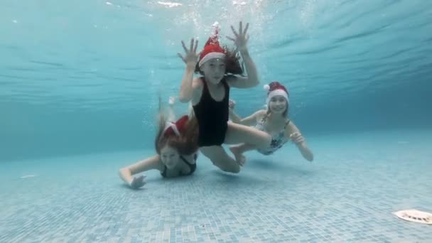 Tres niñas de diferentes edades en los sombreros de Santa celebran la Navidad bajo el agua en la piscina. Se sumergen hasta el fondo de la piscina, miran a la cámara, saludan y sonríen. Retrato. En cámara lenta. 4K . — Vídeos de Stock