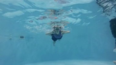 A little girl is engaged in water sports in the pool. She swims underwater in swimming goggles and looks into the camera. Close up. Bottom view. Slow motion. 4K