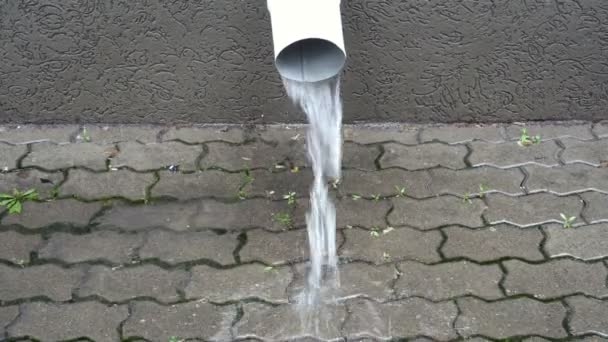 Närbild av ett vitt dräneringsrör på fasaden av byggnaden, varifrån en ström av vatten rinner på trottoaren under regnet. Regn i staden. 4k — Stockvideo