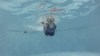 A sporty little girl swims underwater in a pool wearing swimming goggles and looks at the camera. Closeup. Bottom view. Slow motion. 4K