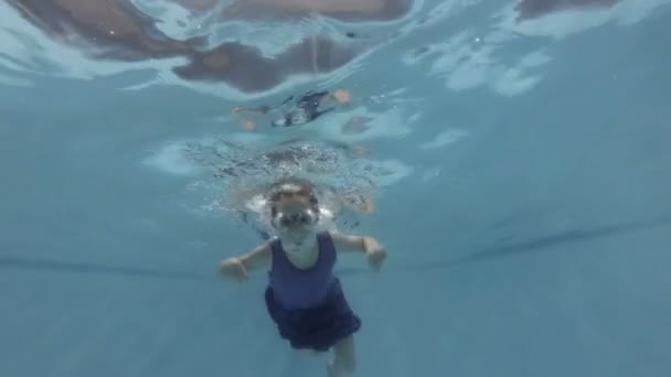 Een kind, een klein meisje, is bezig met duiken. Ze zwemt onder water in het zwembad met een zwembril op, kijkt naar de camera en glimlacht. Sluit maar af. Onderaanzicht. Langzame beweging. 4k — Stockvideo