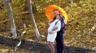 A happy pregnant girl and a man stand under an orange umbrella in a Park on a Sunny autumn day against the backdrop of beautiful nature. They hug, laugh and play. Fashion portrait. 4K