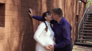 Happy pregnant girl kissing her husband standing near her house on a Sunny day. They laugh, play and together gently stroke the pregnant belly of the girl. Fashion portrait. Concept. 4K