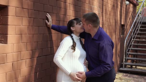 Счастливая беременная девушка целует своего мужа, стоящего рядом с ее домом в солнечный день. Они смеются, играют и вместе нежно гладят беременный живот девушки. Модный портрет. Концепция. 4K — стоковое видео