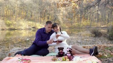 A happy couple, a pregnant girl and a guy, pose sitting in a Park near a pond on a Sunny autumn day with cups of tea in their hands. They hug and smile. Family picnic in nature. 4K
