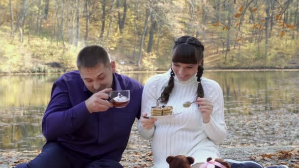 Linda chica embarazada cuchara-alimenta a su marido un pastel sentado cerca de un estanque en el Parque en un día soleado de otoño. Juegan y ríen. Picnic familiar en la naturaleza. Retrato de moda. De cerca. 4K — Vídeo de stock
