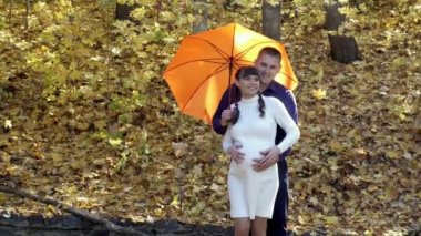 A cute pregnant girl stands with her husband under an orange umbrella in an autumn Park. They hug, play and laugh. A girl in a white knit dress, a guy in a blue shirt. Concept. Closeup. 4K