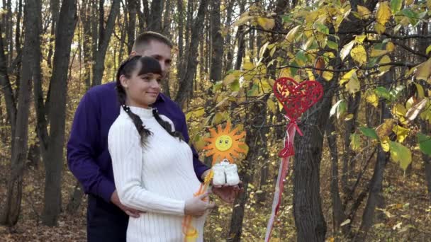 Прекрасная супружеская пара, беременная девушка и парень, позируют в осеннем лесу с белыми детскими носками в руках. Они обнимаются и улыбаются на фоне желтых листьев в солнечный день. 4K — стоковое видео