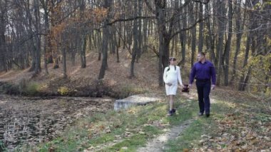 Cute pregnant girl walking with her husband in the autumn Park. They walk, smile, look at each other and hold a soft toy in their hands. A girl in a white knit dress, a guy in a blue shirt. 4K