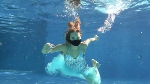 Retrato feminino incomum de uma jovem em uma máscara protetora preta debaixo d 'água. Uma garota mascarada mergulha sob a água na piscina e aperta um dedo de advertência na frente da câmera. Fecha a porta. Movimento lento. — Vídeo de Stock