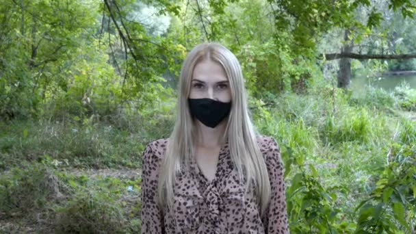 Retrato de una linda chica, rubia, con una máscara protectora negra en la cara, que mira a la cámara, de pie al aire libre sobre un fondo de plantas verdes. Coronavirus. Retrato. Primer plano.. — Vídeo de stock