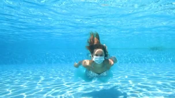 Koncepcyjny portret dziewczyny w masce medycznej pod wodą. Pływa i pozuje do kamery pod wodą w pustym basenie. Koronawirus. Covid-19. Druga fala. Zbliżenie. Zwolnij trochę. 4K. — Wideo stockowe