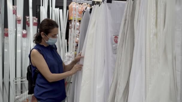 Une femme adulte célibataire dans un masque médical examine et évalue les rideaux blancs dans un atelier vide avant d'acheter. Concept. Coronavirus. Épidémie. Deuxième vague. Distance sociale. Gros plan. 4K. — Video