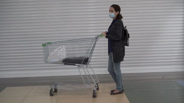 Tıbbi maskeli yetişkin bir kadın alışveriş arabasıyla bir mağazada beyaz bir duvarın önünde duruyor ve etrafına bakıyor. Sosyal mesafeyi koruyor. Kavram. Coronavirus. Salgın hastalık. İkinci dalga. Genel plan. — Stok video
