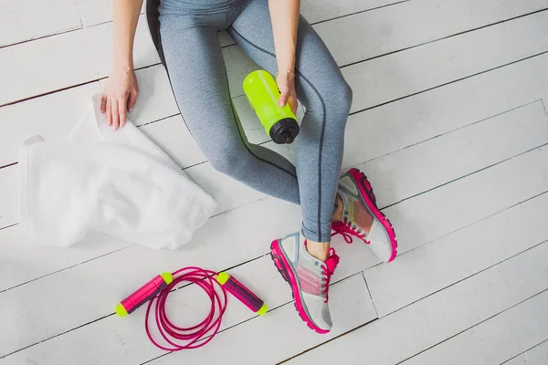 Athletic skinny girl in sport wear and sneakers sitting on the wooden floor in the gym and holding a bottle in her hands. Sport concept.