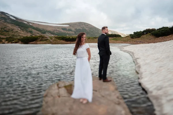 Wedding concept photo shoot. Beautiful young couple of groom and bride in white wedding dress standing on a big stone in a lake in spring mountains with snow