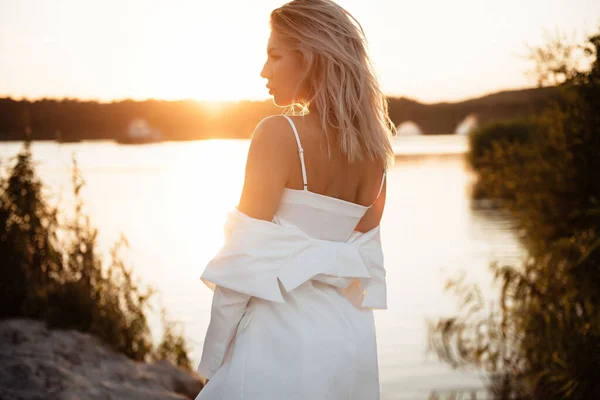 Side view of gorgeous blonde in white suit standing near lake during summer sunset. Young woman posing on camera among beautiful nature.