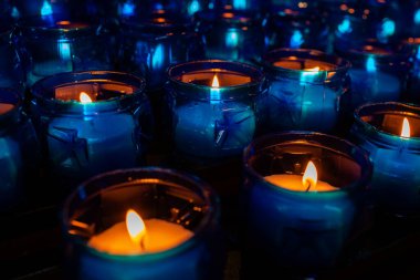 Rows of blue glass candles with glowing flames create a peaceful ambiance, casting warm light and reflections in the surrounding dark space, enhancing the serene environment