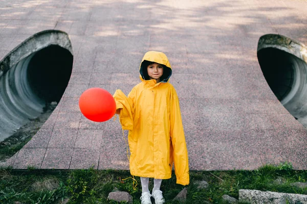 little girl in oversized yellow raincoat with red balloon against sewage tunnel pipe