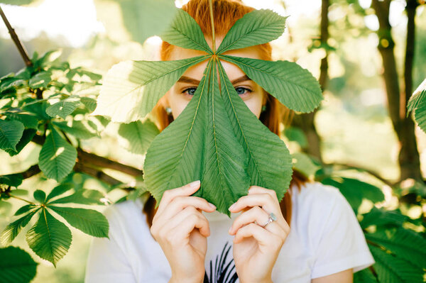 obscured view of young red hair woman looking at camera behind chestnut leaf in park