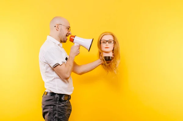 bald man with loudspeaker holding plastic mannequin head on yellow backdrop
