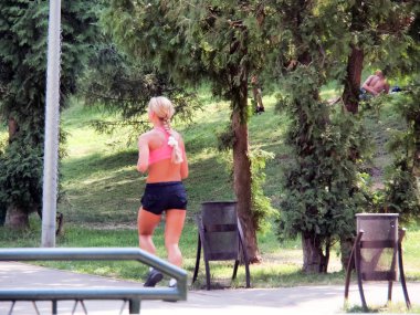 Bir adam ağaçların arasındaki çimlerin üzerinde yatıyor. Aşağıda, bayan bir atlet boş teneke vazoların önünden geçiyor. Tembelliğini bir kenara bırak..
