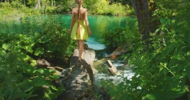 Uzun örgülü bir kadın ormanda açık bir derenin üzerinde yalınayak yürüyor..