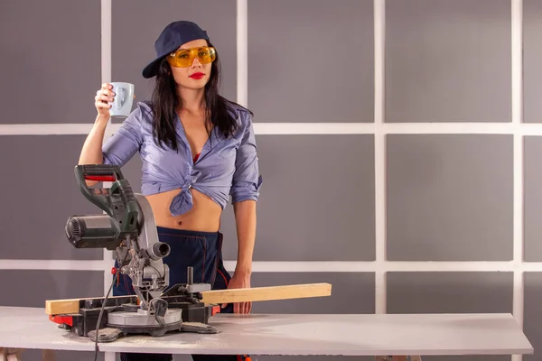 Mavi gömlekli mutlu bir genç kadın atölyede alet ve güvenlik ekipmanları dolu çalışma tezgahında fincan ile ayakta kamera bakarak ve gülümseyerek .
