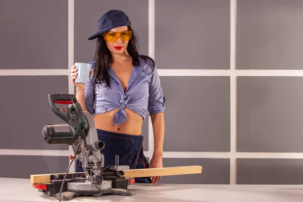 Mavi gömlekli mutlu bir genç kadın atölyede alet ve güvenlik ekipmanları dolu çalışma tezgahında fincan ile ayakta kamera bakarak ve gülümseyerek .