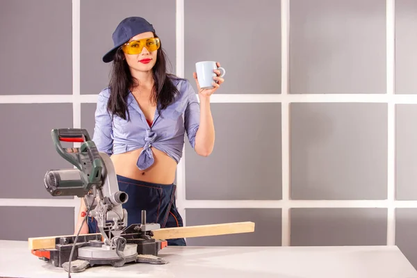 Mavi gömlekli mutlu bir genç kadın atölyede alet ve güvenlik ekipmanları dolu çalışma tezgahında fincan ile ayakta kamera bakarak ve gülümseyerek .