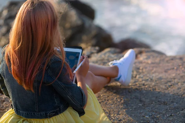 Piękne blond dziewczyna siedzi na skale nad morzem i rysuje na tablecie. Styl lato wakacje. Nowoczesna dziewczyna — Zdjęcie stockowe