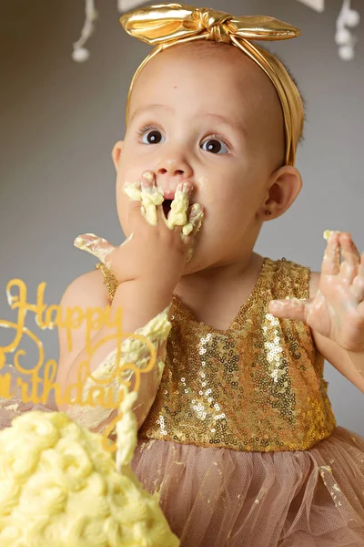 Pequeña niña dulce en un vestido de oro con un lazo en la cabeza tratando de un pastel de gelatina jazzy de una crema. estudio toma de un cumpleaños sobre un fondo gris rodeado de bolas —  Fotos de Stock